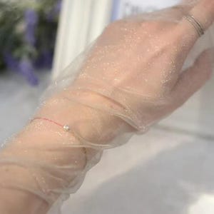 May include: A sheer, light beige glove with a subtle shimmer. A delicate gold bracelet with a small, clear gem is visible on the wrist. A silver ring is on a finger. The glove is slightly translucent.
