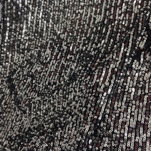 May include: A close-up of a black fabric covered in silver and black sequins. The sequins are arranged in a pattern of vertical lines.