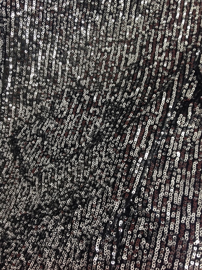 May include: A close-up of a black fabric covered in silver and black sequins. The sequins are arranged in a pattern of vertical lines.