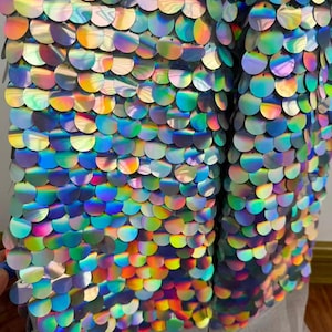 May include: Close-up of a fabric covered in iridescent, circular sequins. The sequins are arranged in a scale-like pattern, reflecting a rainbow of colors including blue, green, yellow, and purple. The fabric appears to be layered over a black mesh.