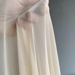 May include: A close-up of a piece of off-white mesh fabric. The fabric is very sheer and has a fine, even weave.