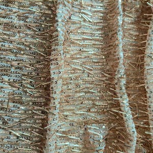 May include: A sheer fabric featuring a dense arrangement of metallic gold sequins and thin, elongated gold bead accents. The embellishments are arranged in horizontal rows across the light-colored, semi-transparent mesh base material.