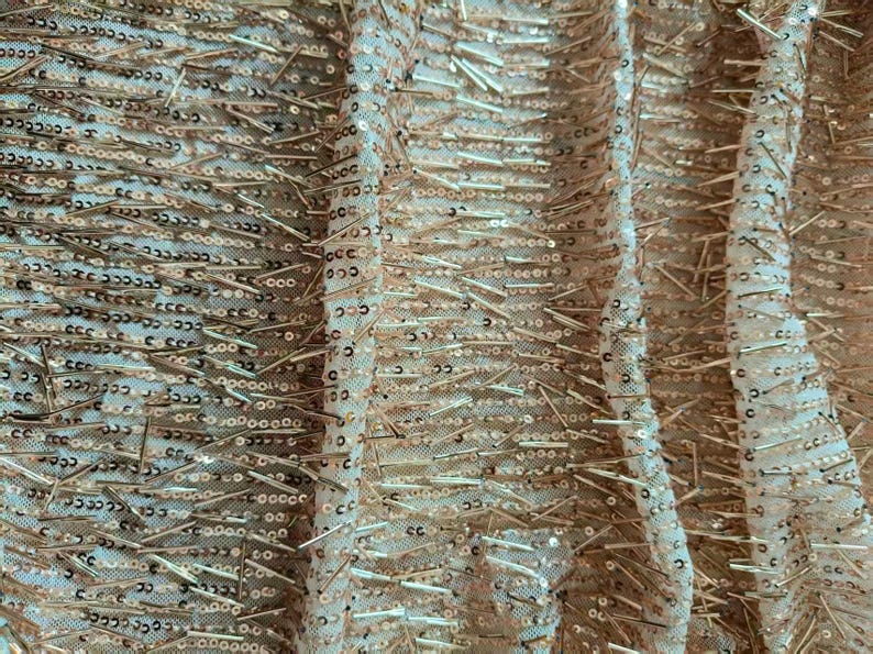 May include: A sheer fabric featuring a dense arrangement of metallic gold sequins and thin, elongated gold bead accents. The embellishments are arranged in horizontal rows across the light-colored, semi-transparent mesh base material.