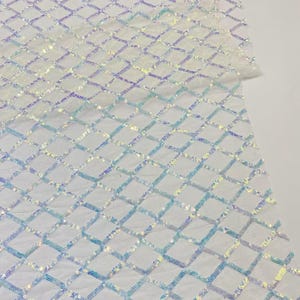 Peut inclure: Tissu irisé à motif losange. Le tissu blanc est orné d'une grille de paillettes chatoyantes dans les tons de bleu, de violet et d'argent. Les paillettes captent la lumière, créant un effet scintillant.