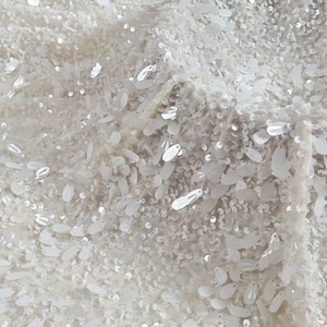 May include: Close-up of white fabric with dense embellishments. The fabric is covered in sequins and iridescent, leaf-shaped beads, creating a shimmering effect. Suitable for bridal or evening wear.