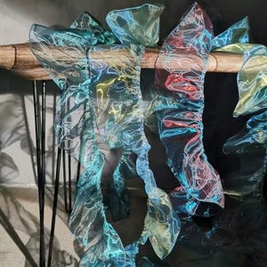 May include: Iridescent fabric streamers in shades of teal, blue, and red hang from a wooden beam. The sheer material has a shimmering, almost holographic quality, with white vein-like patterns. The streamers are gathered and ruffled, creating a flowing, decorative effect.