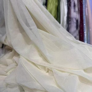 May include: Close-up of ivory-colored fabric with a delicate, sheer texture. The fabric is draped and folded, creating soft shadows and highlights. The material appears lightweight and flowing, suitable for apparel or decorative purposes.