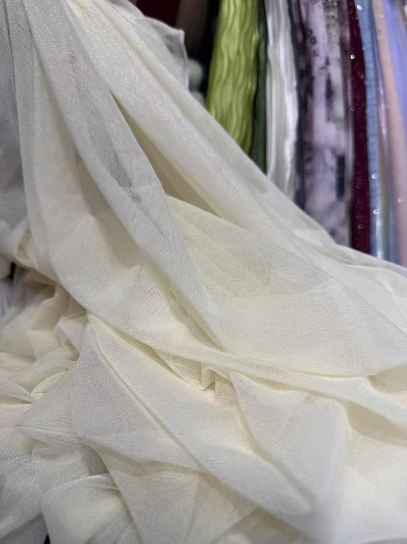 May include: Close-up of ivory-colored fabric with a delicate, sheer texture. The fabric is draped and folded, creating soft shadows and highlights. The material appears lightweight and flowing, suitable for apparel or decorative purposes.