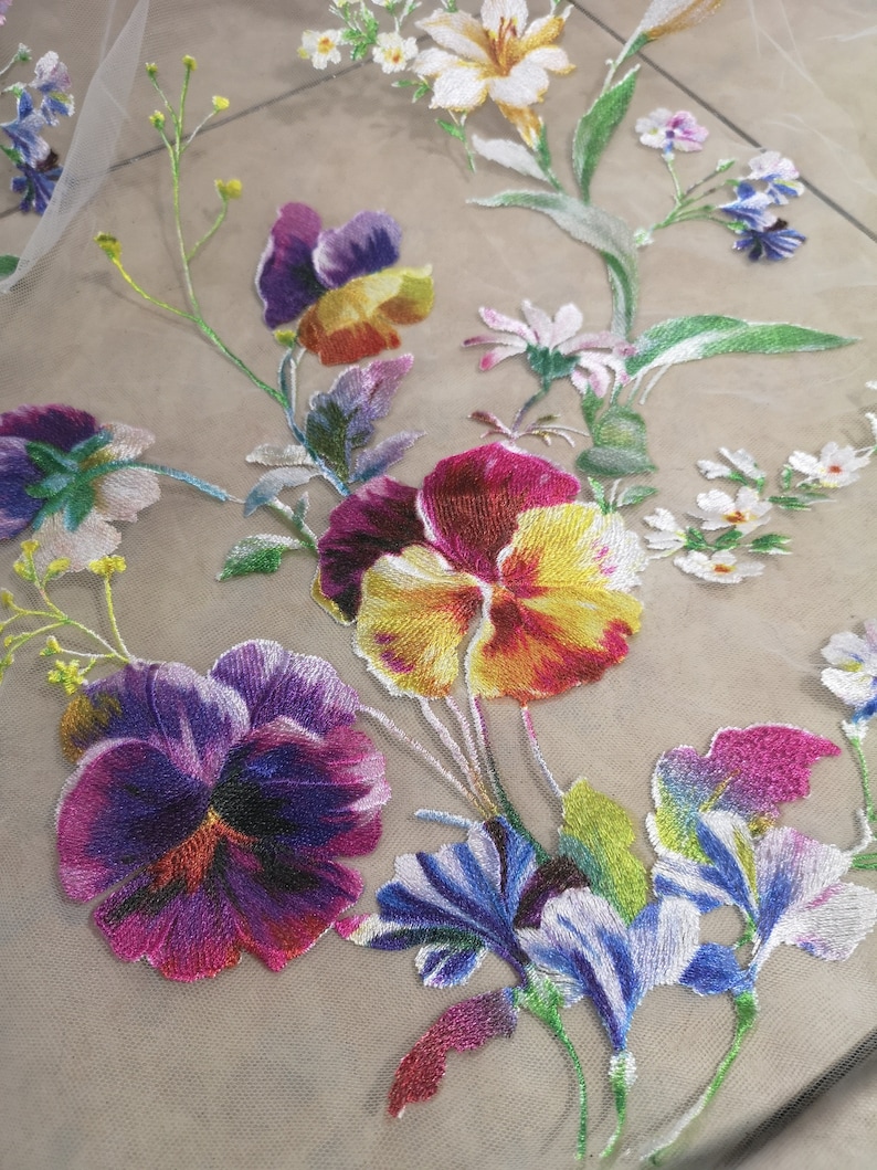 May include: Embroidered floral fabric featuring a range of colourful flowers, including purple, yellow, red, and blue. The fabric is sheer and delicate, and the flowers are intricately detailed.