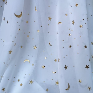 May include: A sheer white fabric featuring a repeating pattern of metallic gold crescent moons and stars of varying sizes scattered across the surface. The material is draped to create soft folds, highlighting the reflective gold foil print.