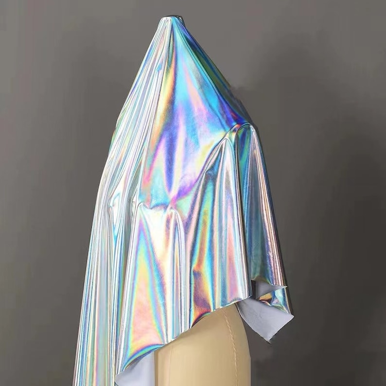 May include: A silver iridescent fabric with a holographic finish. The fabric is draped over a form and has a smooth, shiny texture.