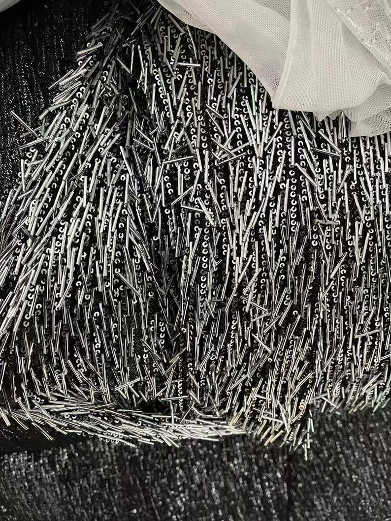 K&ouml;nnte beinhalten: Nahaufnahme eines schwarzen Stoffes mit silbernen Pailletten und Fransen. Die Fransen bestehen aus kleinen, d&uuml;nnen, silbernen St&auml;ben.