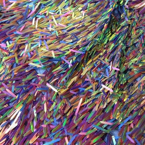 Puede incluir: Un primer plano de una tela iridiscente con lentejuelas largas, delgadas y coloridas. Las lentejuelas están dispuestas en un patrón aleatorio y crean un efecto brillante. La tela es de color azul claro y tiene una textura suave y sedosa.