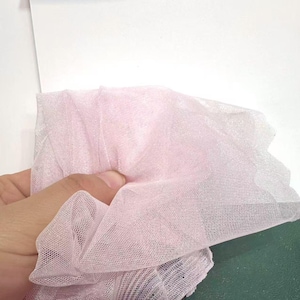 May include: A close-up of a piece of light pink tulle fabric. The fabric is held in a hand and is bunched up, showing its delicate, sheer texture. The tulle is a soft, pastel color, and the weave is fine and open.
