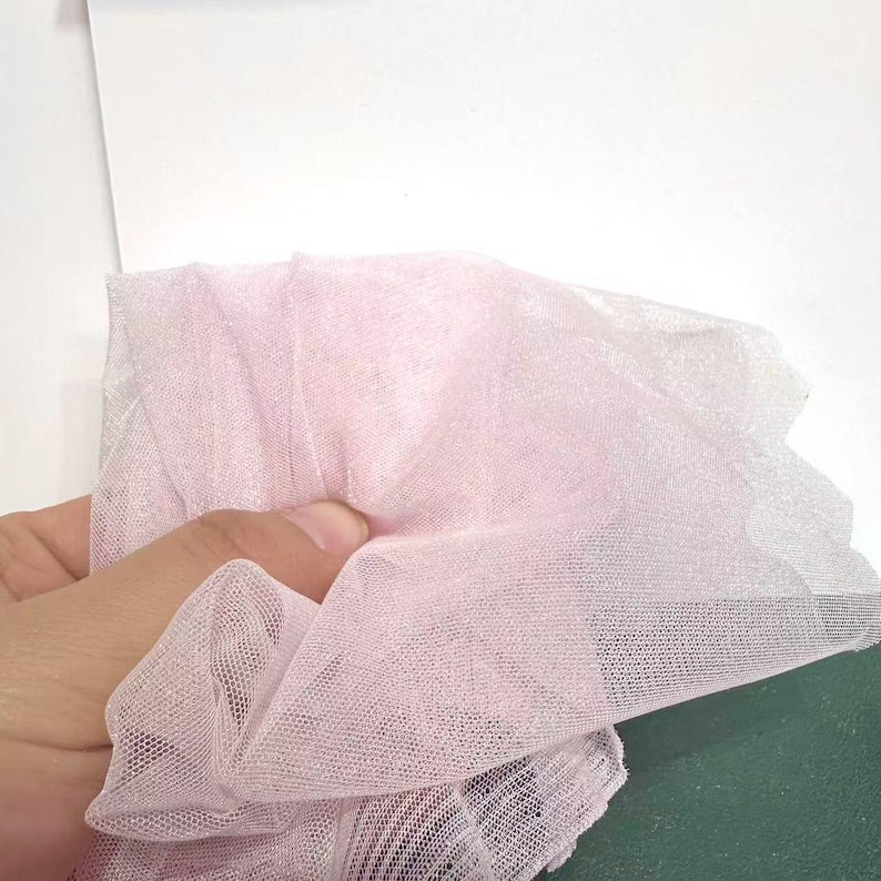 May include: A close-up of a piece of light pink tulle fabric. The fabric is held in a hand and is bunched up, showing its delicate, sheer texture. The tulle is a soft, pastel color, and the weave is fine and open.