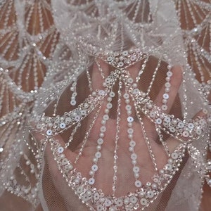 May include: Close-up of white, sheer fabric with intricate beaded and sequined embellishments. The design features a fan-like pattern with rows of small, round beads and larger sequins, creating a shimmering effect. The fabric is held up, showing the delicate texture.