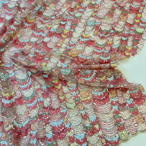 May include: A close-up view of a decorative fabric with a repeating, layered, scalloped pattern. The fabric is a mix of red, white, blue, yellow, and green, with a shimmering, sequined texture. The design is ornate and eye-catching.