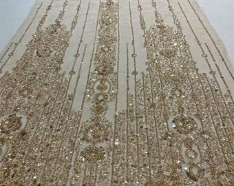 May include: Ivory fabric embellished with intricate gold beading and sequins. The design features ornate patterns with vertical and circular elements. The fabric is suitable for bridal wear, evening gowns, or other special occasion garments.