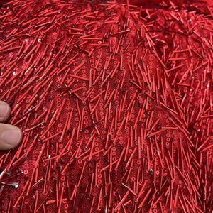 K&ouml;nnte beinhalten: Roter Stoff mit einer strukturierten Oberfl&auml;che, bedeckt mit kleinen roten Pailletten und langen, d&uuml;nnen roten Perlen.
