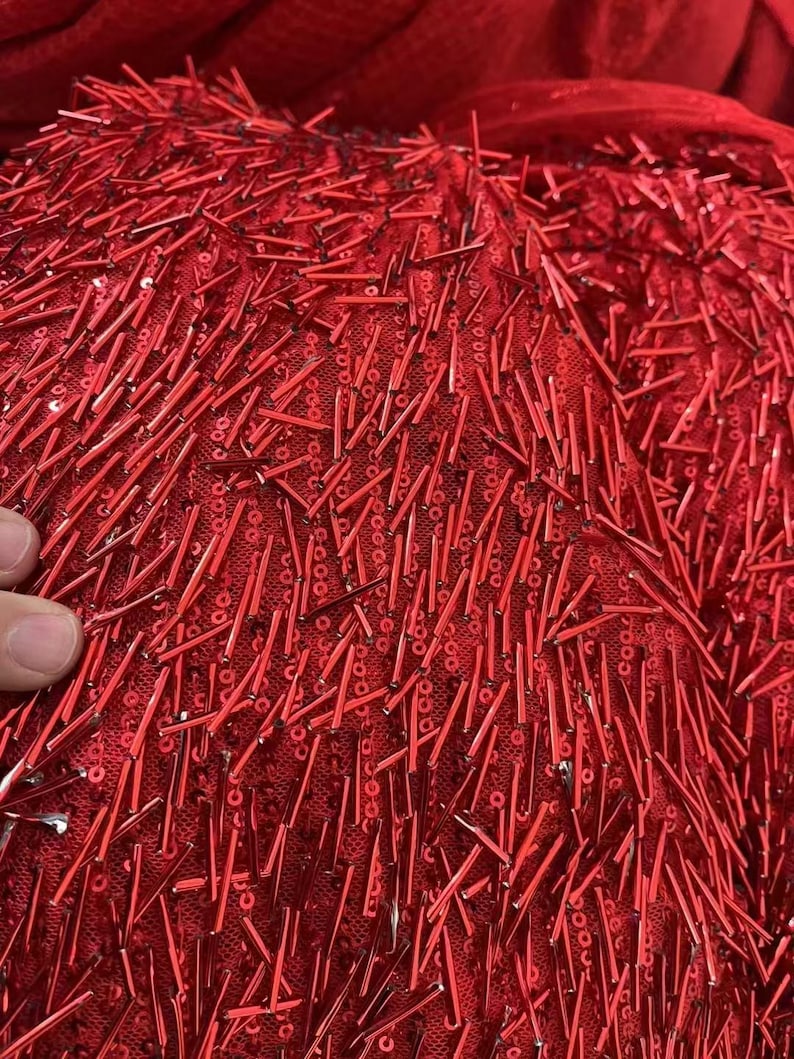 K&ouml;nnte beinhalten: Roter Stoff mit einer strukturierten Oberfl&auml;che, bedeckt mit kleinen roten Pailletten und langen, d&uuml;nnen roten Perlen.