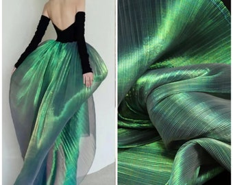 Può includere: Tessuto plissettato verde iridescente con un leggero luccichio. Il tessuto è leggero e trasparente, con un drappeggio morbido e fluido.