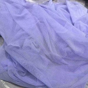 May include: A close-up of a lavender-colored tulle fabric. The fabric is draped and folded, creating a textured appearance. The sheer material allows light to pass through, highlighting the delicate weave and subtle sheen.