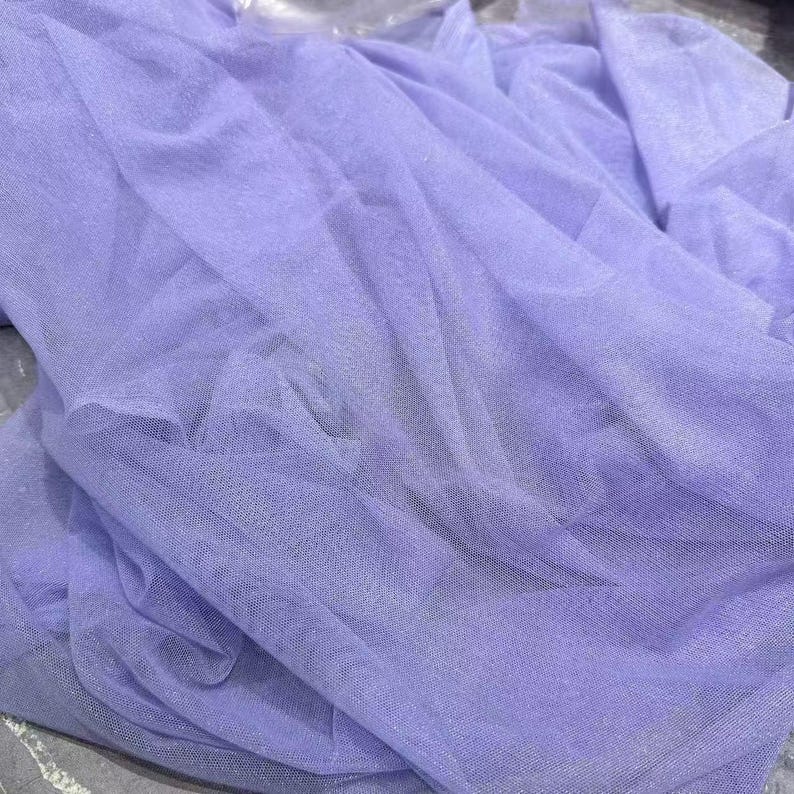 May include: A close-up of a lavender-colored tulle fabric. The fabric is draped and folded, creating a textured appearance. The sheer material allows light to pass through, highlighting the delicate weave and subtle sheen.
