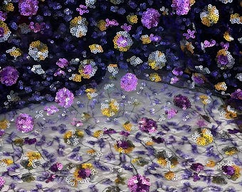 May include: A sheer black mesh fabric featuring an intricate pattern of embroidered floral motifs. The design is heavily embellished with shimmering sequins in vibrant shades of purple, gold, and silver, creating a dense, textured surface.