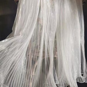 May include: White sheer fabric with a shimmery finish. The fabric is draped over a chair and has a pleated texture.