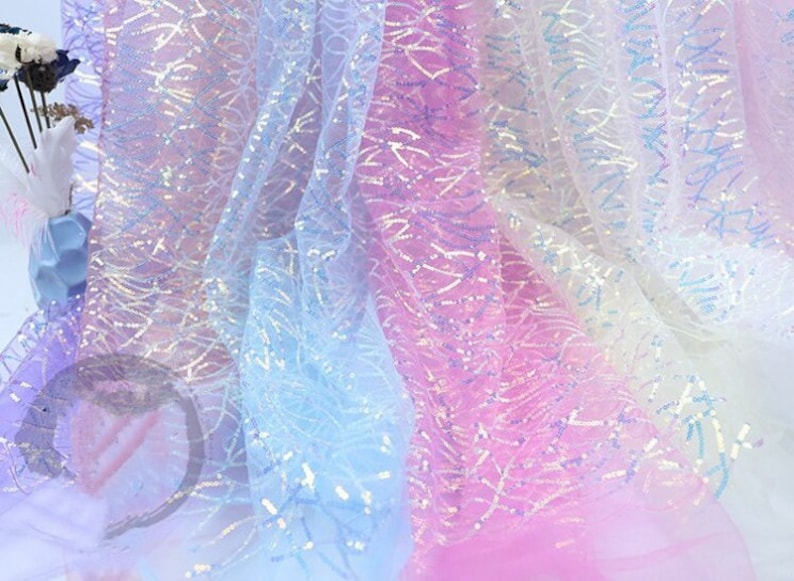 May include: A close-up of iridescent fabric with a delicate, patterned design. The fabric is layered in shades of light blue, pink, and white, creating a soft and ethereal look.