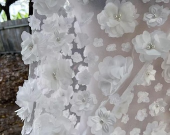 May include: Close-up of white fabric adorned with 3D floral embellishments. The flowers vary in size and are accented with small, silver-colored beads. The fabric appears to be a sheer material, creating a delicate and textured look.