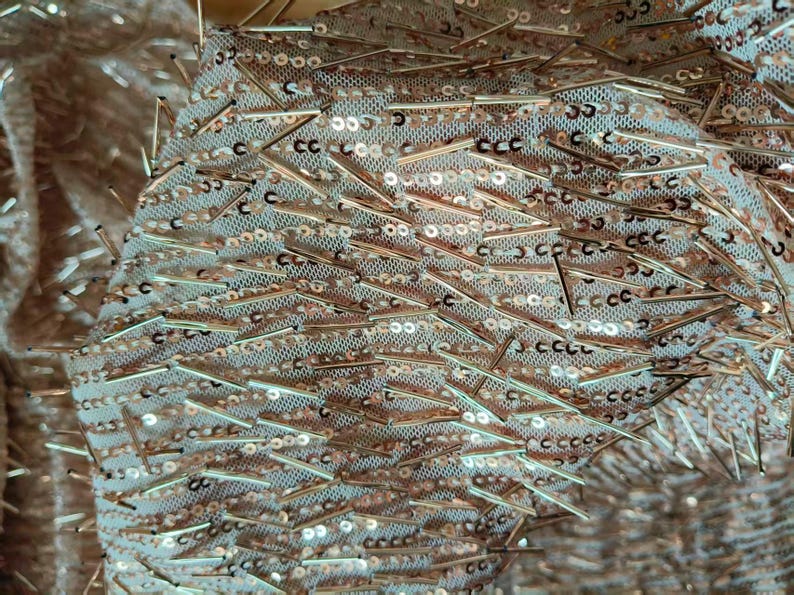 May include: A sheer fabric base in a light champagne tone, densely embellished with metallic rose gold sequins and thin, elongated rose gold bugle beads arranged in a scattered, multi-directional pattern across the entire surface.