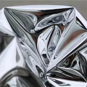 Könnte beinhalten: Ein zerknittertes Blatt aus glänzendem, silberfarbenem Material. Das Material ist stark reflektierend und erzeugt abstrakte Muster aus Licht und Schatten.