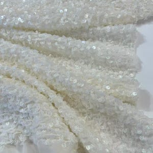 Peut inclure: Gros plan sur un tissu blanc recouvert de petites paillettes scintillantes. Le tissu est plié, créant une surface texturée avec des profondeurs et des reflets variables. Les paillettes captent la lumière, créant un effet scintillant.