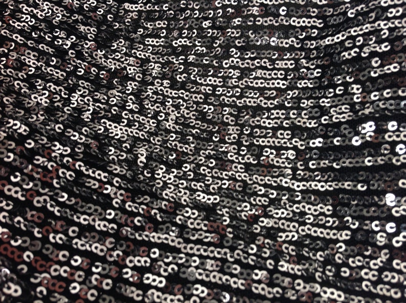 May include: Close-up of a black fabric covered in silver sequins. The sequins are arranged in a repeating pattern.