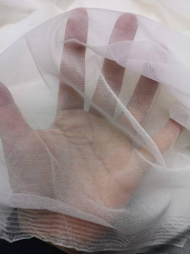 May include: Close-up of sheer white fabric with a fine mesh texture. The fabric is layered, creating depth and highlighting the transparency. The material appears soft and delicate, ideal for crafting or decorative purposes.