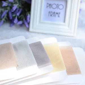 May include: A selection of sheer, neutral-toned fabric swatches, including beige, gray, and yellow, are displayed on white cards. A white photo frame with the text "PHOTO FRAME" is in the background, along with purple flowers.