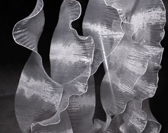 Può includere: Un lungo runner da tavola bianco e trasparente caratterizzato da una texture molto arricciata e ondulata. Il tessuto è traslucido con un sottile luccichio, creando una serie di pieghe verticali e sinuose che scendono dal bordo di una superficie in legno.