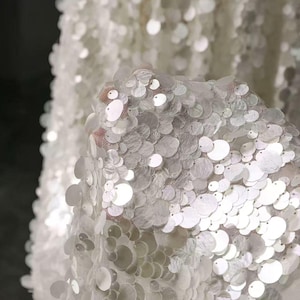 Puede incluir: Una base de tela blanca transparente densamente cubierta con lentejuelas blancas circulares superpuestas. Las lentejuelas son reflectantes y varían ligeramente en tamaño, creando una superficie texturizada y brillante que capta la luz.