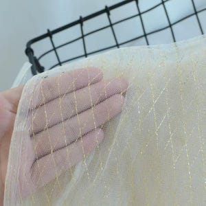 May include: A sheer, off-white tulle fabric featuring a delicate, diagonal grid pattern woven with shimmering gold metallic thread. The lightweight, semi-transparent material has a fine mesh texture that allows light to pass through clearly.