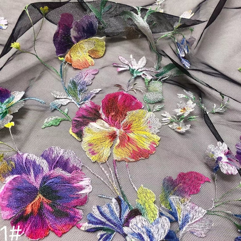 May include: Black sheer fabric with embroidered floral designs. The flowers are in shades of purple, pink, yellow, and green. The fabric has a delicate, airy feel.