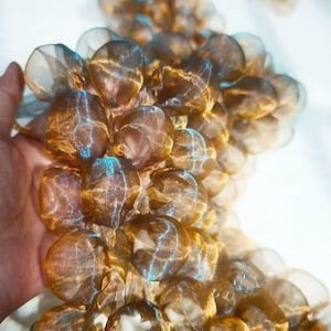 Peut inclure: Gros plan sur un amas d'orbes translucides brun doré, possiblement en tissu ou éléments décoratifs. Les orbes ont une texture délicate en forme de toile et sont tenus dans une main sur fond blanc. Les orbes ont une légère teinte bleue.