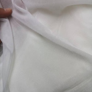 May include: Close-up of white sheer fabric, possibly tulle or organza. The fabric is folded and draped, showing its delicate texture and translucence. The material appears soft and lightweight, ideal for crafting or apparel.
