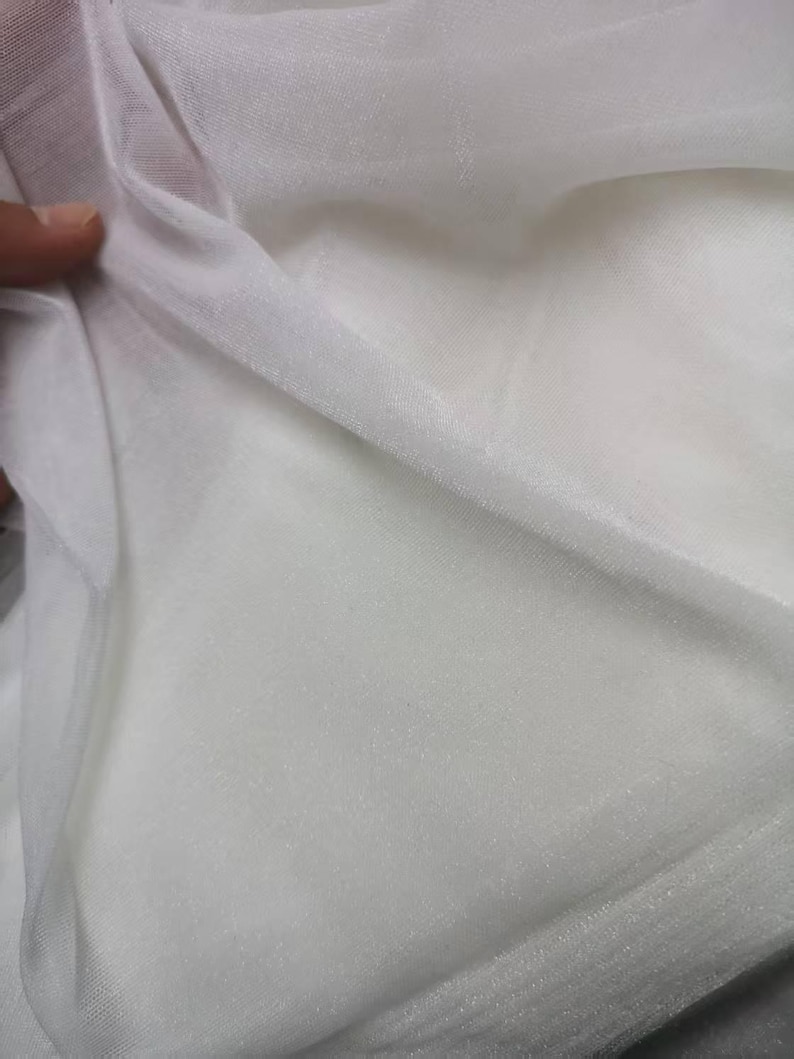 May include: Close-up of white sheer fabric, possibly tulle or organza. The fabric is folded and draped, showing its delicate texture and translucence. The material appears soft and lightweight, ideal for crafting or apparel.