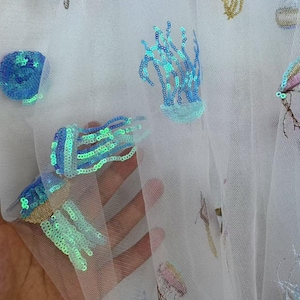 Puede incluir: Tela blanca transparente con criaturas marinas bordadas y con lentejuelas. El diseño incluye medusas, corales y otras criaturas marinas azules, verdes y doradas. La tela se sostiene, mostrando los detalles del bordado.