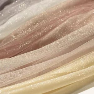 May include: Close-up of shimmering fabric in a gradient of colors. The fabric transitions from a dark grey to white, then to a light pink, and finally to a pale yellow. The material has a subtle sparkle.