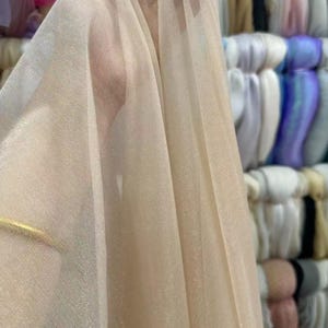 May include: Close-up of a sheer, beige fabric with a fine mesh texture. The fabric is draped, revealing folds and subtle variations in color. The background shows stacks of colorful textiles.