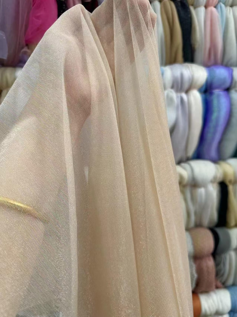May include: Close-up of a sheer, beige fabric with a fine mesh texture. The fabric is draped, revealing folds and subtle variations in color. The background shows stacks of colorful textiles.