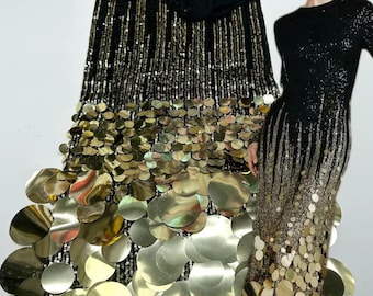 May include: A black fabric featuring a dense pattern of vertical gold sequins that transition into large, reflective gold circular discs at the bottom. The fabric is displayed alongside a long-sleeved black dress with the same sequin and disc design.