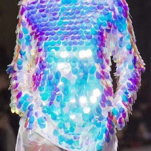 Puede incluir: Una blusa de lentejuelas iridiscentes de manga larga con un esquema de color azul, morado y rosa. Las lentejuelas están dispuestas en un patrón de escamas.